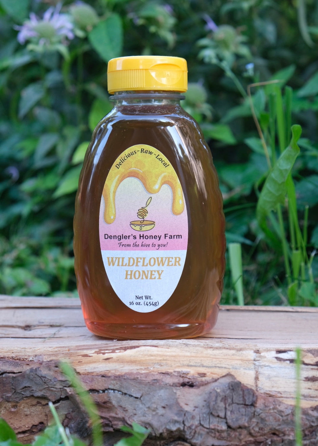 A bottle of Dangler's Honey Farm 16-ounce Wildflower Honey sits on a rustic wooden log with a lush green garden background.